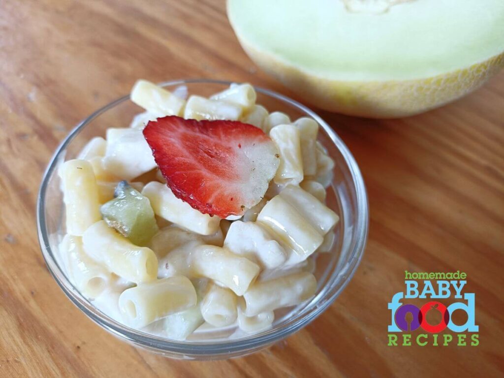 A bowl of fruit and macaroni salad next to a melon wedge